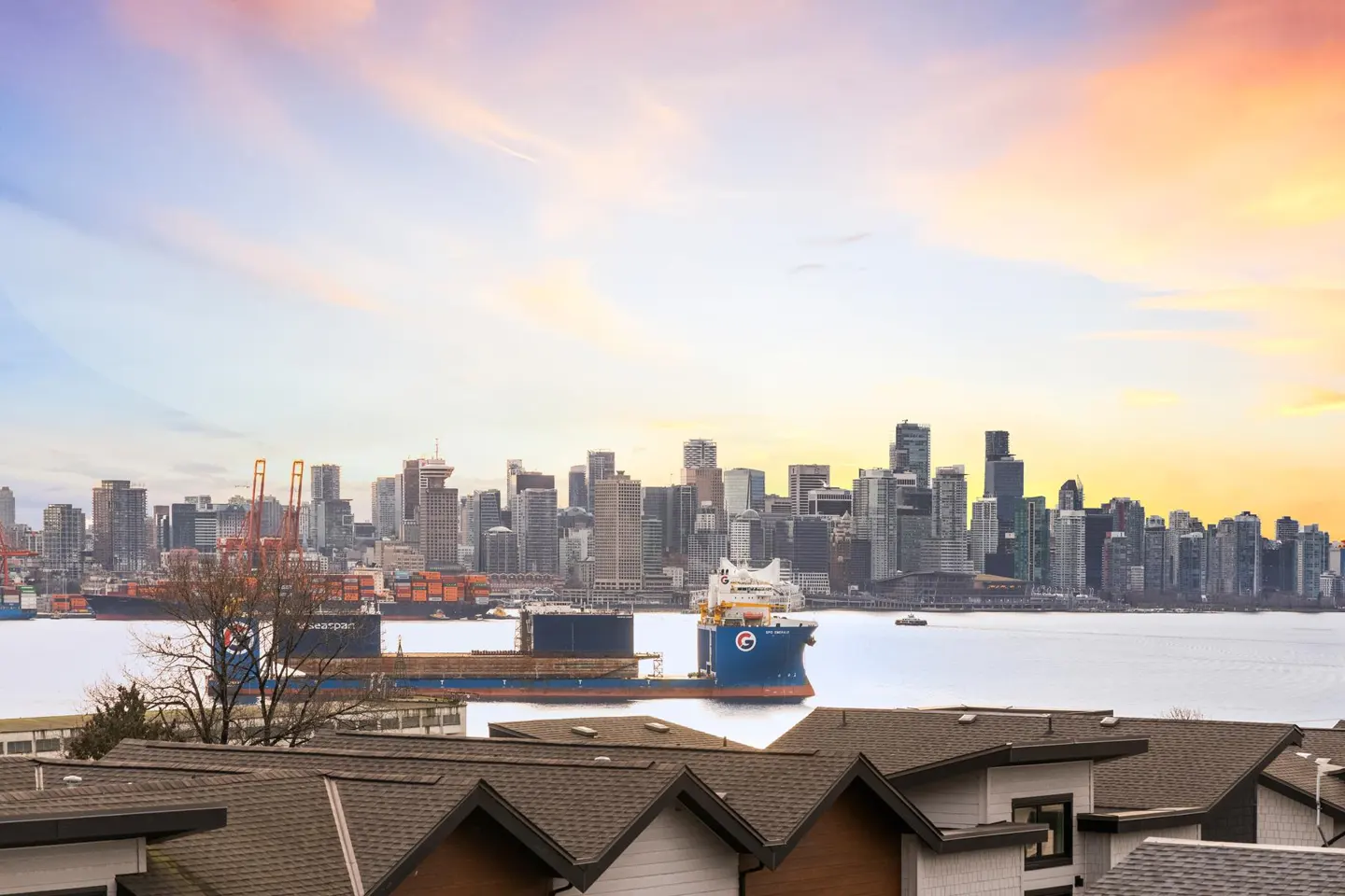 View of the Vancouver skyline and harbor with a large cargo ship at sunset, seen from above residential rooftops.