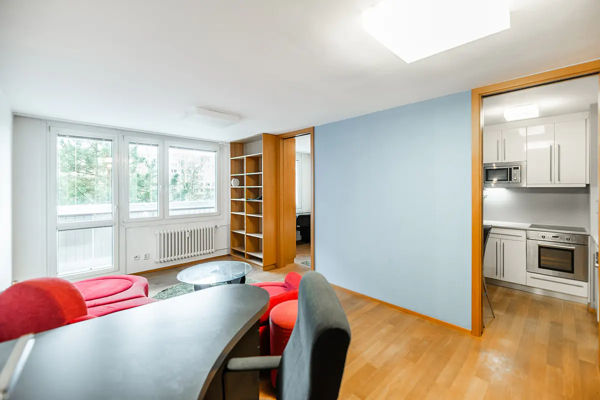 Bright apartment interior with wood floors, blue wall, red seating, and a view into a white kitchen.