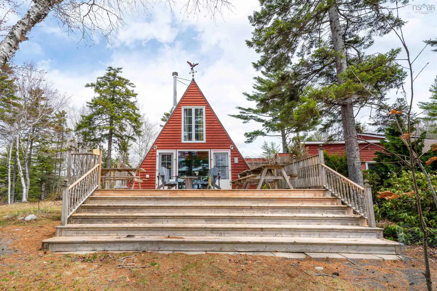 A-frame red house with a large deck and wooden stairs, surrounded by trees.