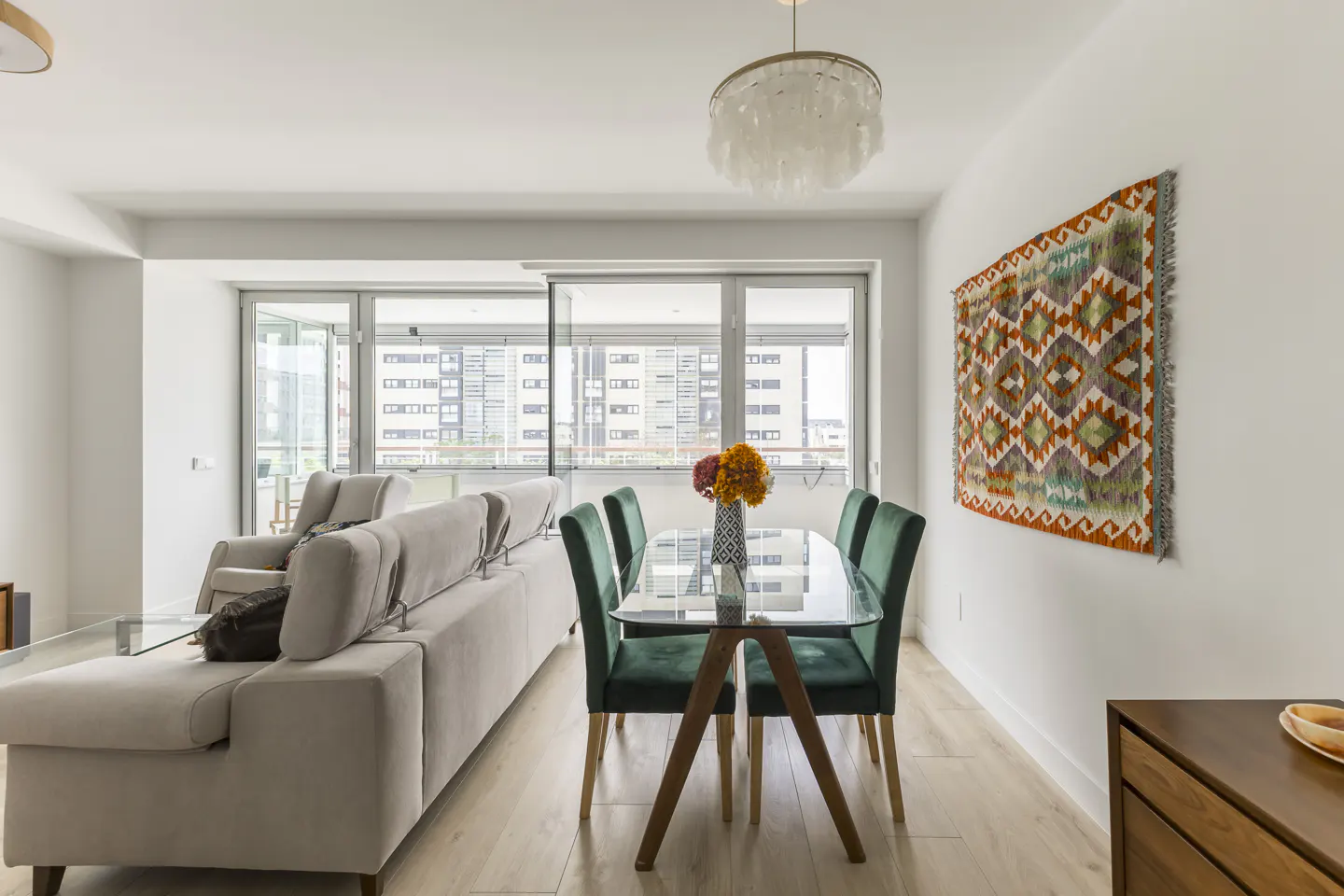 Bright, modern living space with a beige sofa, glass dining table with green chairs, and a colorful patterned wall hanging.