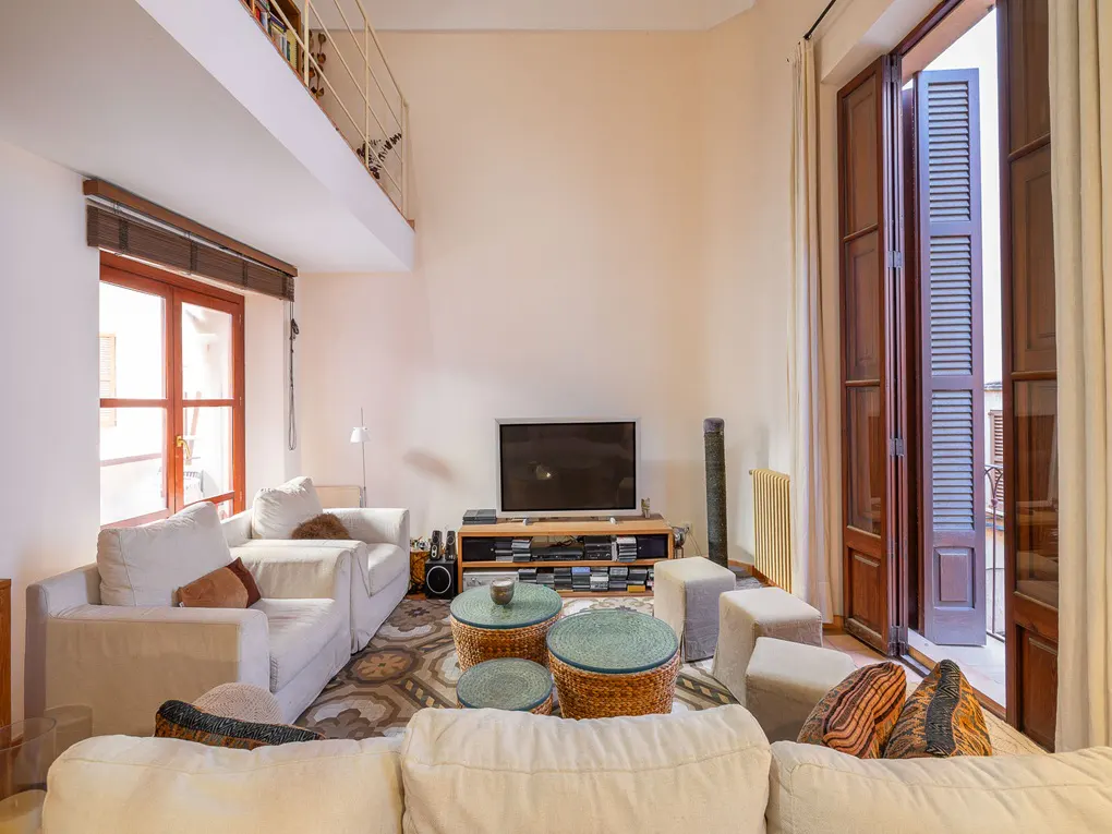 Bright living room with white sofas, woven tables, and a TV. Balcony doors are open, letting in light. A loft area is visible above.