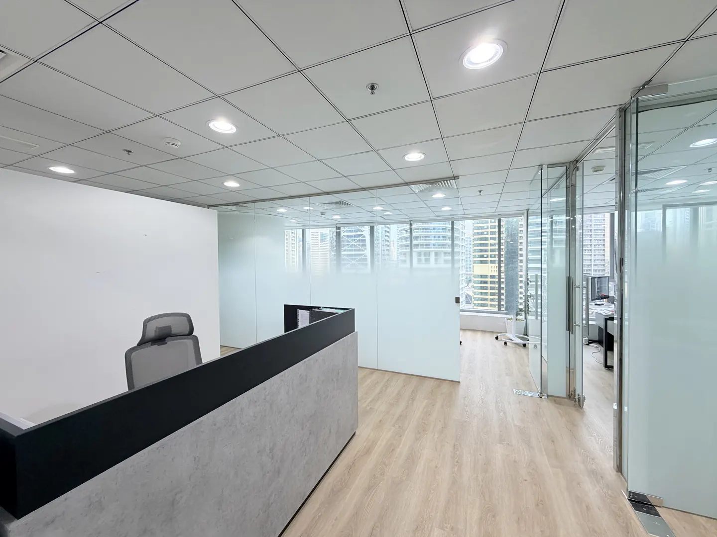 Bright office space with wood floors, frosted glass walls, and a gray reception desk. City buildings are visible through the windows.