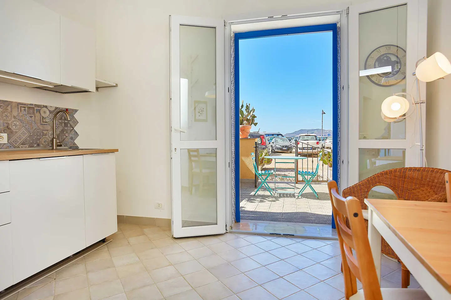 Bright apartment interior with white cabinets, a dining table, and an open door to a patio with a view of parked cars and the sea.