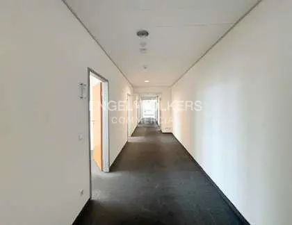 A long, empty hallway with white walls and a dark gray floor. Doors are visible on the left side.