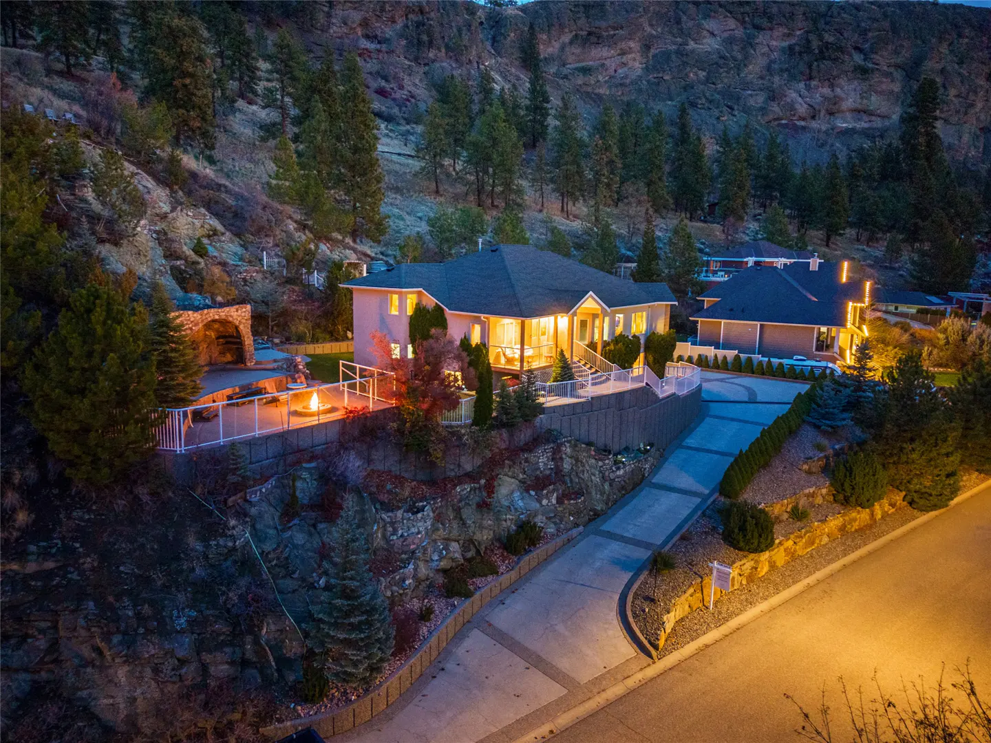 A dusk view of a beige house with a dark roof, nestled into a rocky hillside with pine trees. A fire pit glows on a patio.