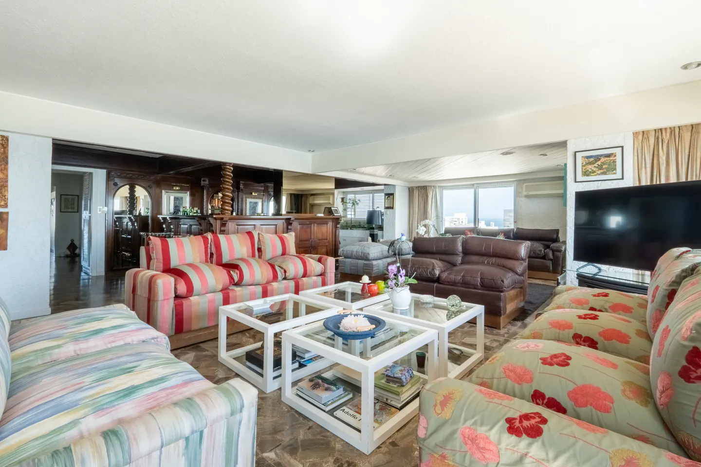A spacious living room with colorful sofas, glass coffee tables, and a built-in bar area. Large windows offer a city view.