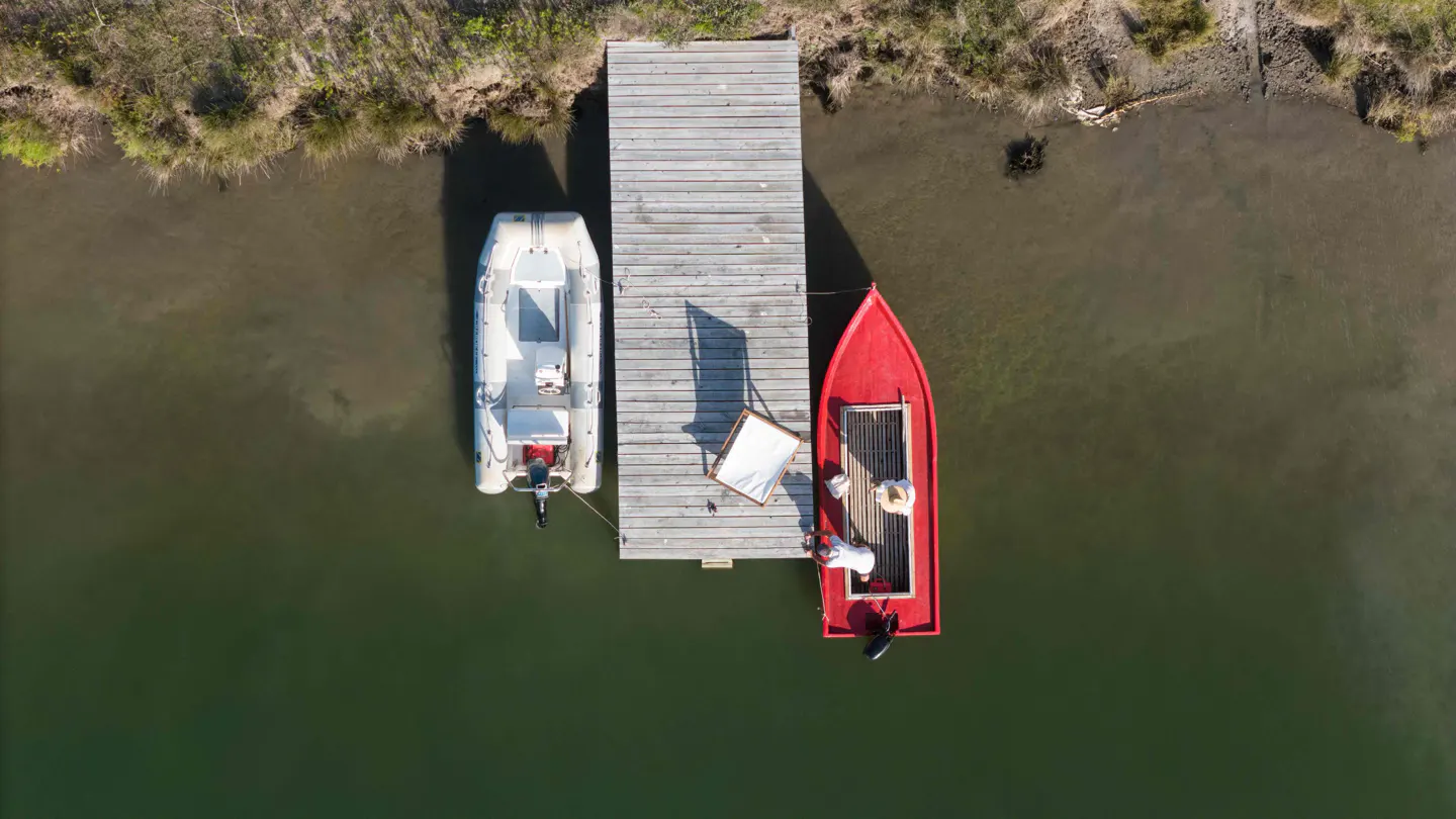 Aerial view of a wooden dock with a white inflatable boat and a red boat docked on either side. Two people are on the red boat.