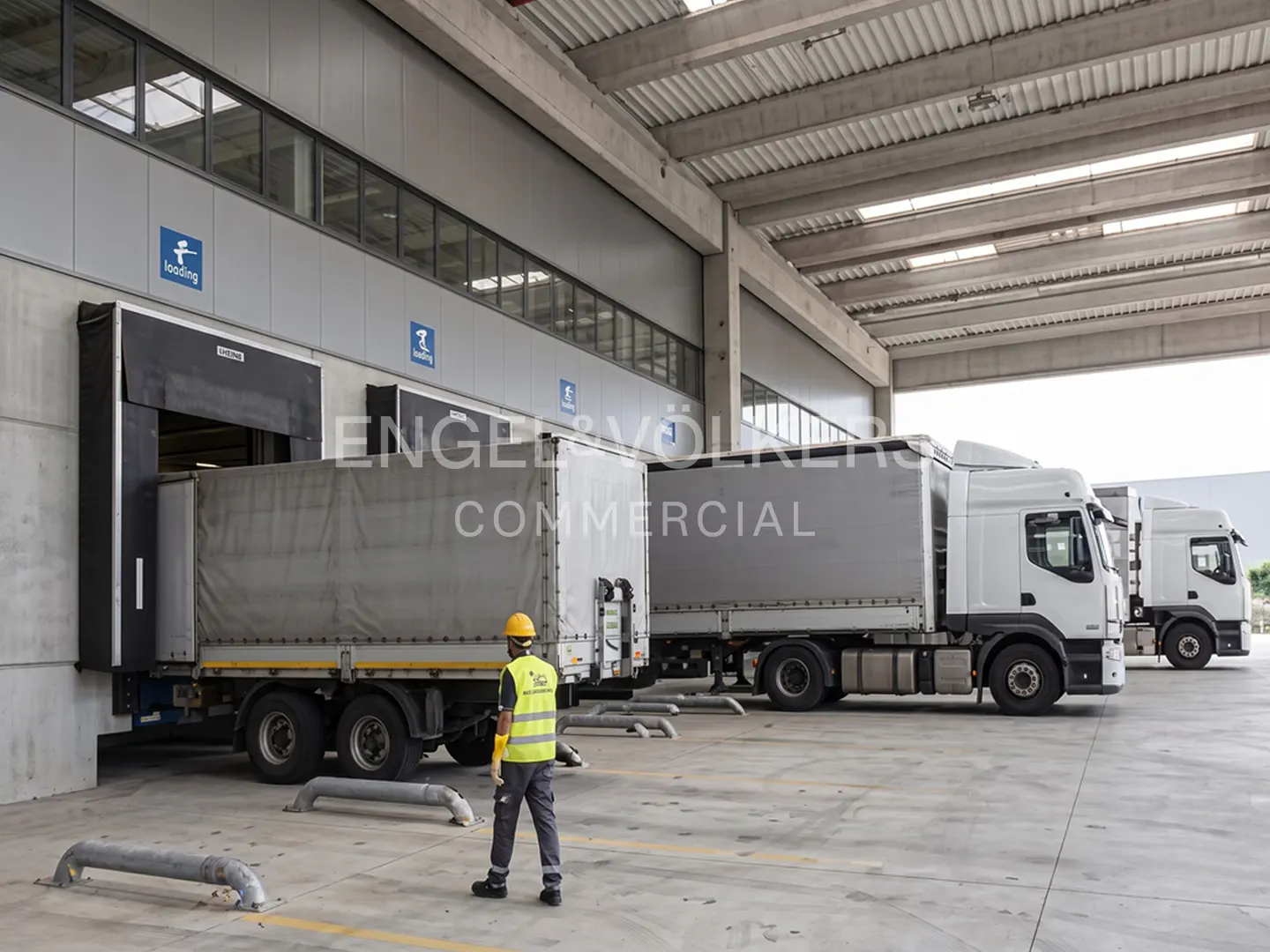 A worker in a yellow vest stands near trucks at a loading dock. The trucks are white with gray trailers.