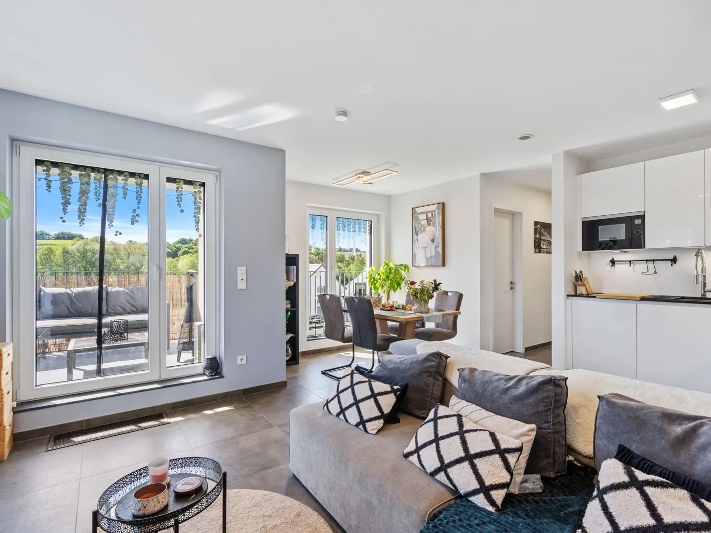 A bright, modern apartment interior with a gray sofa, dining table, and white kitchen. Balcony doors open to a green view.