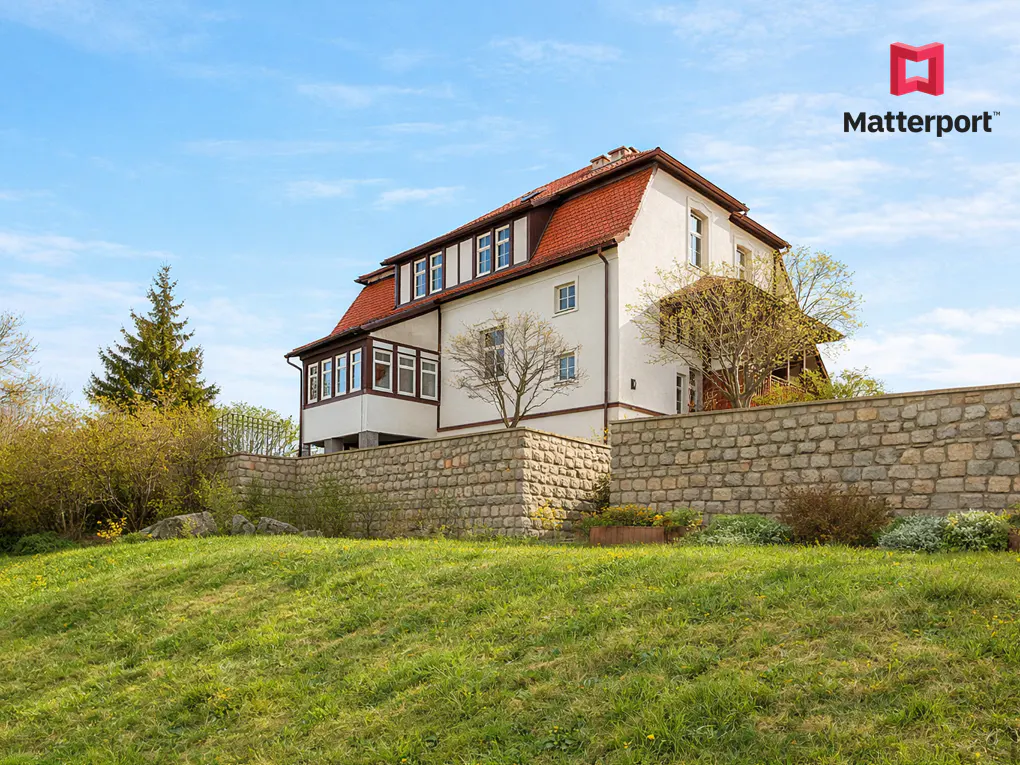 Exterior view of a two-story house with a red roof, white walls, and a stone wall in front. Green lawn and blue sky.
