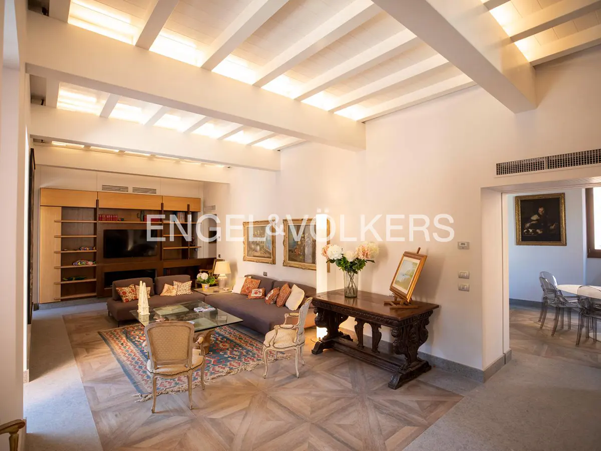 Spacious living room with white beamed ceilings, grey sofas, patterned rug, and an ornate wooden console table.