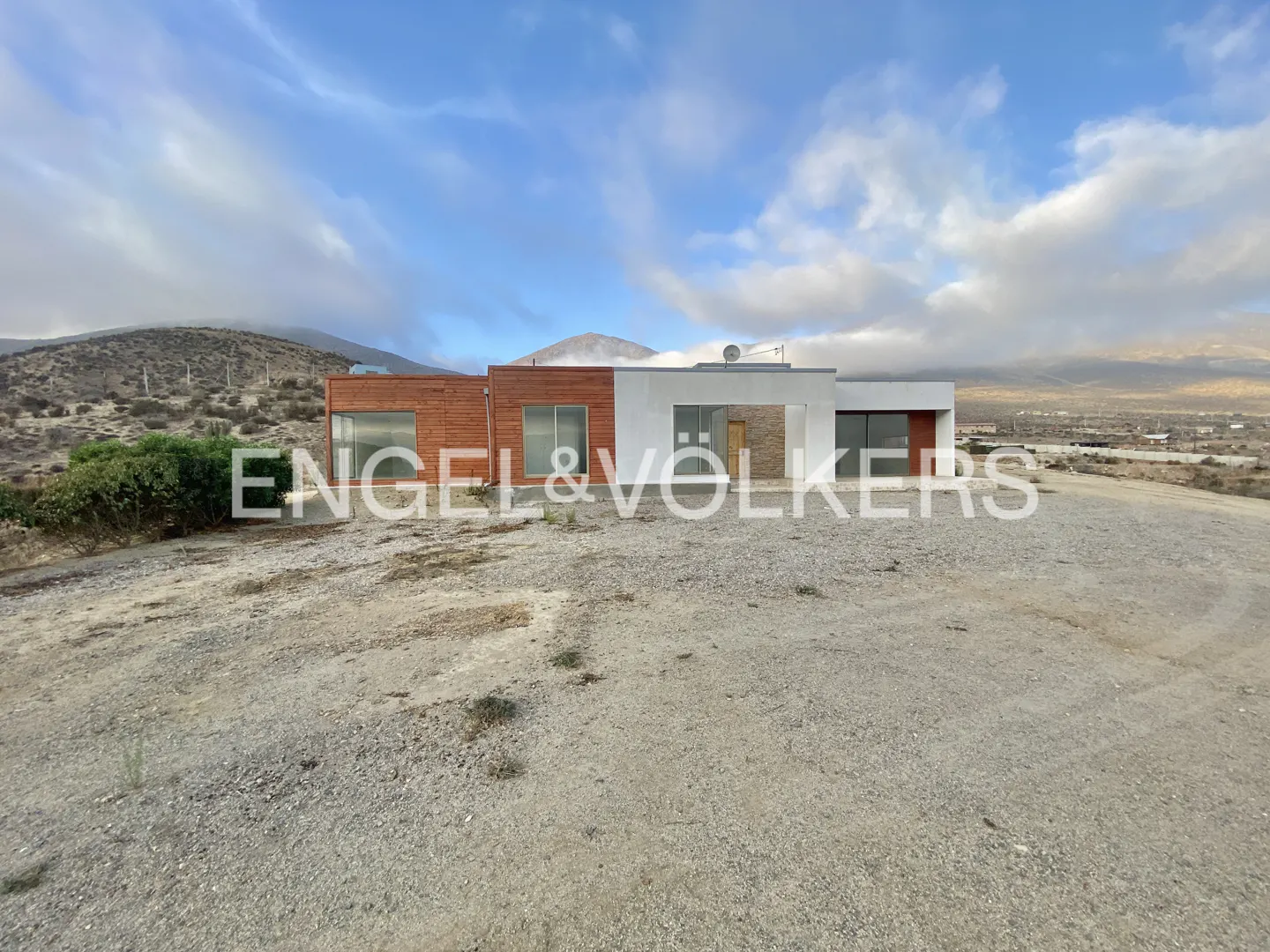 Modern home with wood and white siding on a gravel lot under a cloudy sky. Engel & Volkers logo is superimposed.