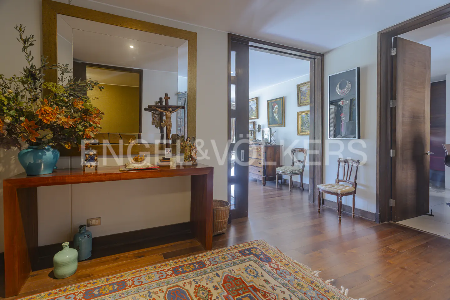 Entryway with a wooden console table, a blue vase with flowers, and a large mirror. A doorway leads to a room with paintings and furniture.