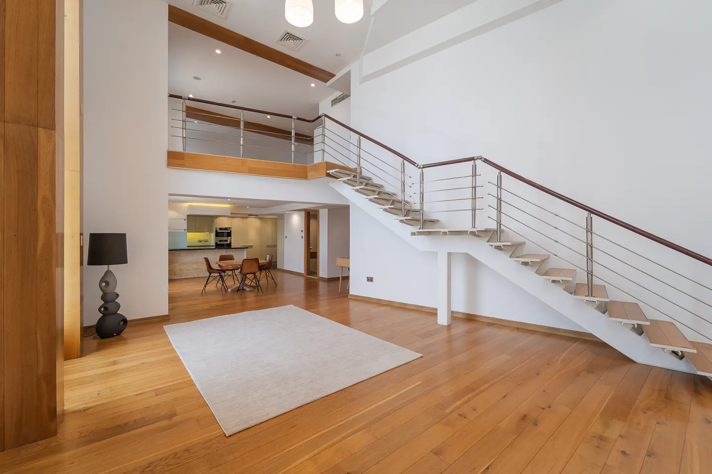 Bright, modern living room with wood floors, white walls, and a staircase with metal railings. A dining area and kitchen are visible in the background.