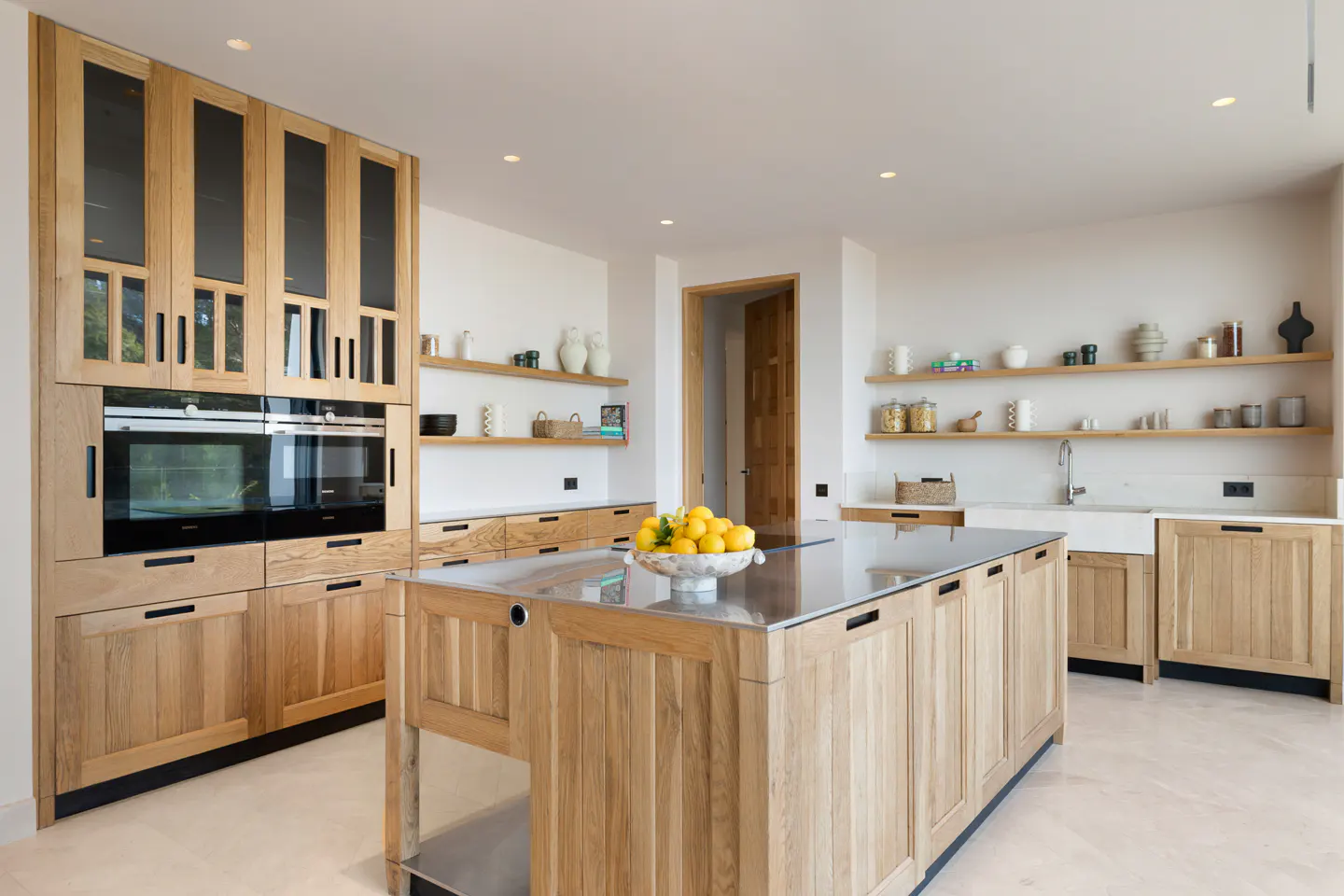 Bright kitchen with light wood cabinets, black accents, and a stainless steel island topped with a bowl of lemons. Open shelves display decorative items.