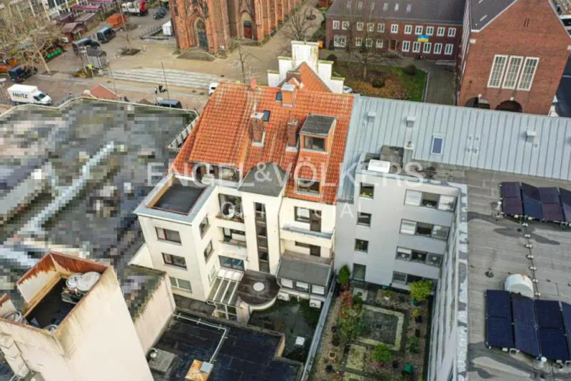 Aerial view of a white building with a red tile roof, surrounded by other buildings and a church in the background.