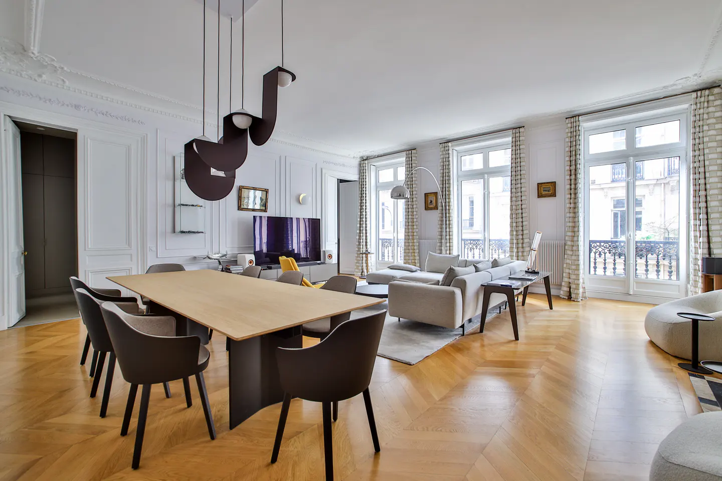 Modern living room with herringbone wood floors, a light wood dining table, dark chairs, and a white sectional sofa.