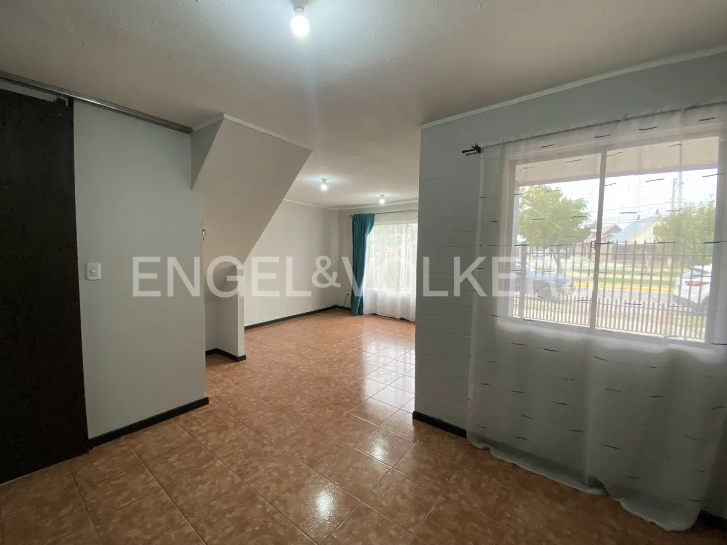 A bright, empty living room with brown tile floors, white walls, and a large window with sheer curtains.