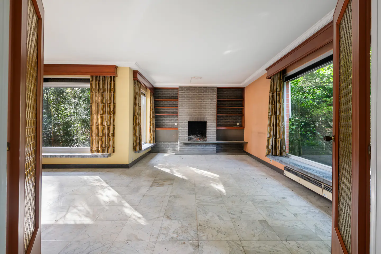A bright, empty living room with marble floors, a brick fireplace, and large windows with patterned curtains.