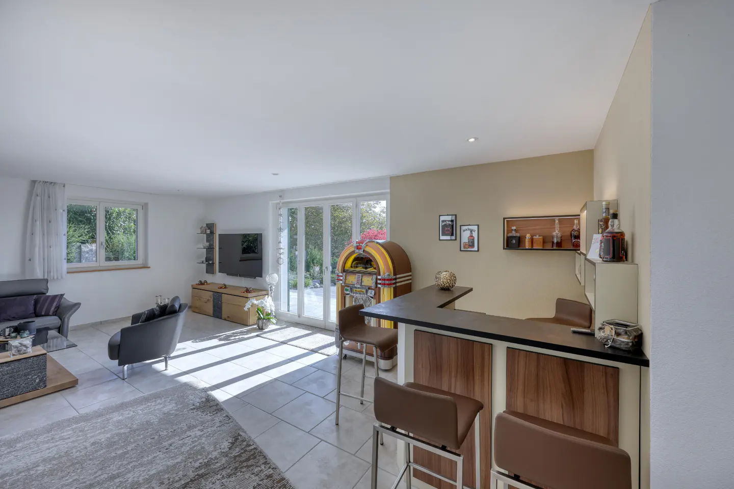 Bright living room with a bar, jukebox, and seating area. Sunlight streams through windows and doors.