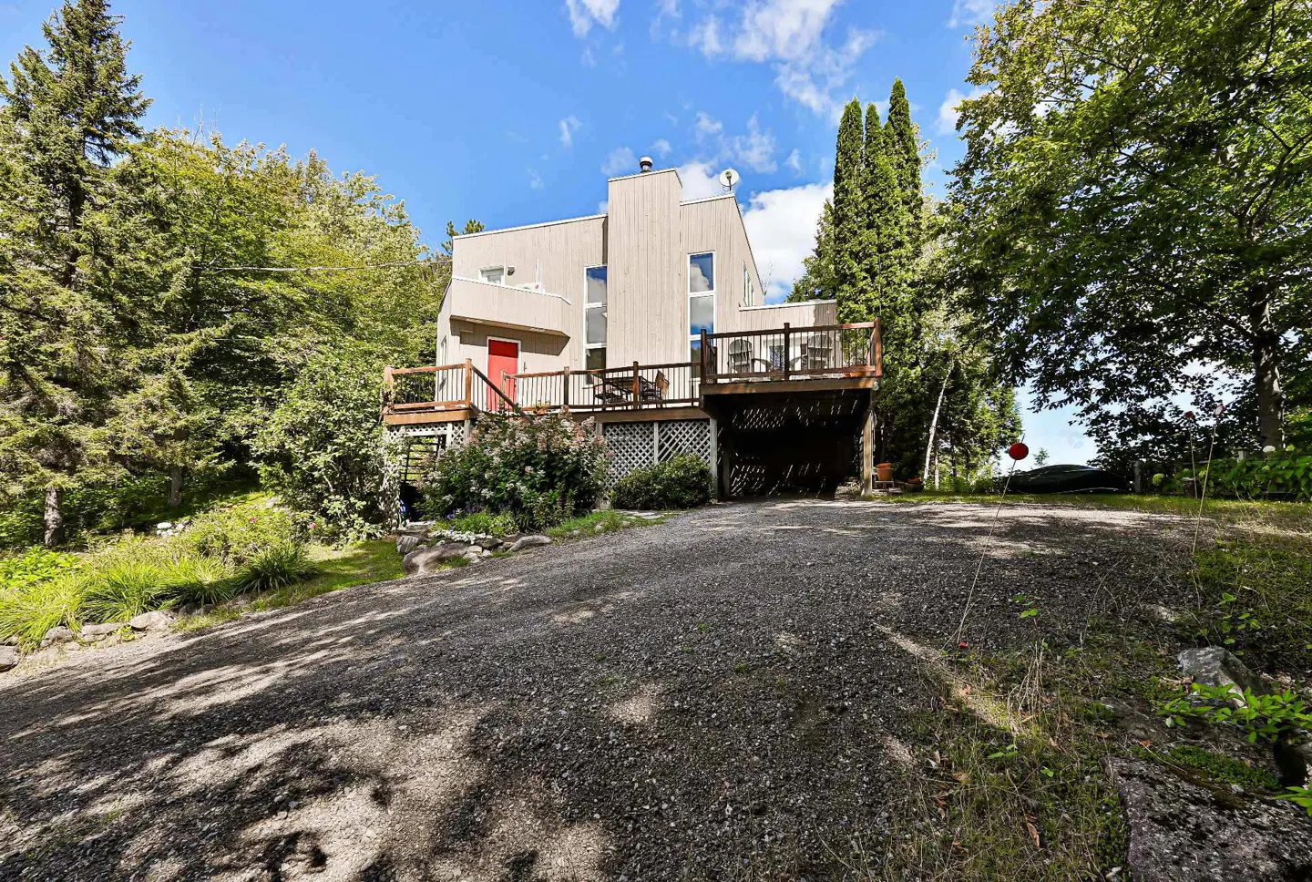 Exterior view of a modern beige house with a red door and a wooden deck, surrounded by trees.