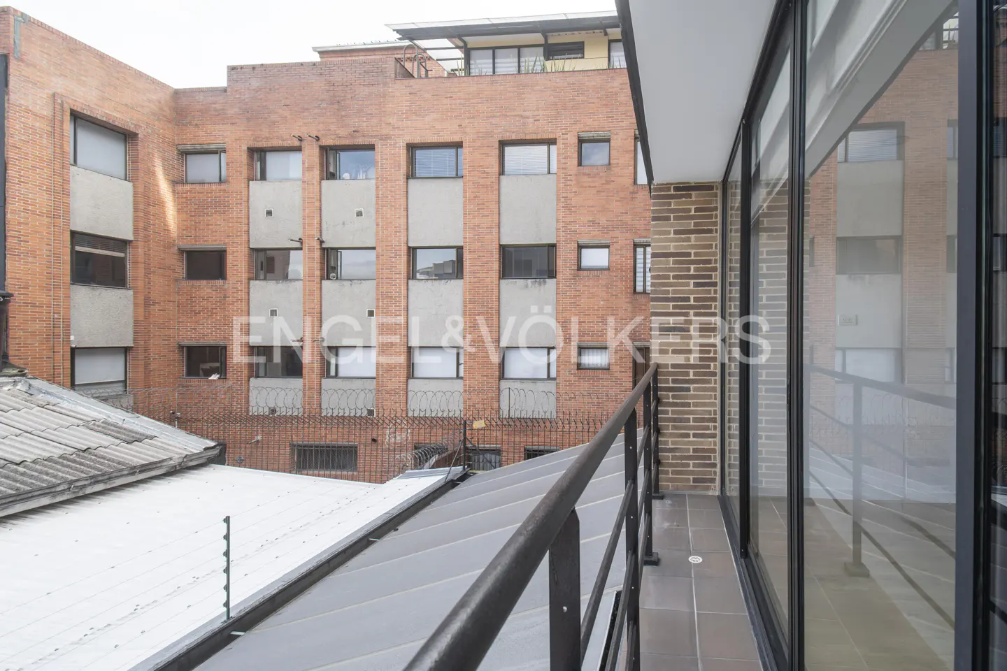 Balcony view of a brick building with windows, a gray roof, and a black railing. The balcony has large glass windows.