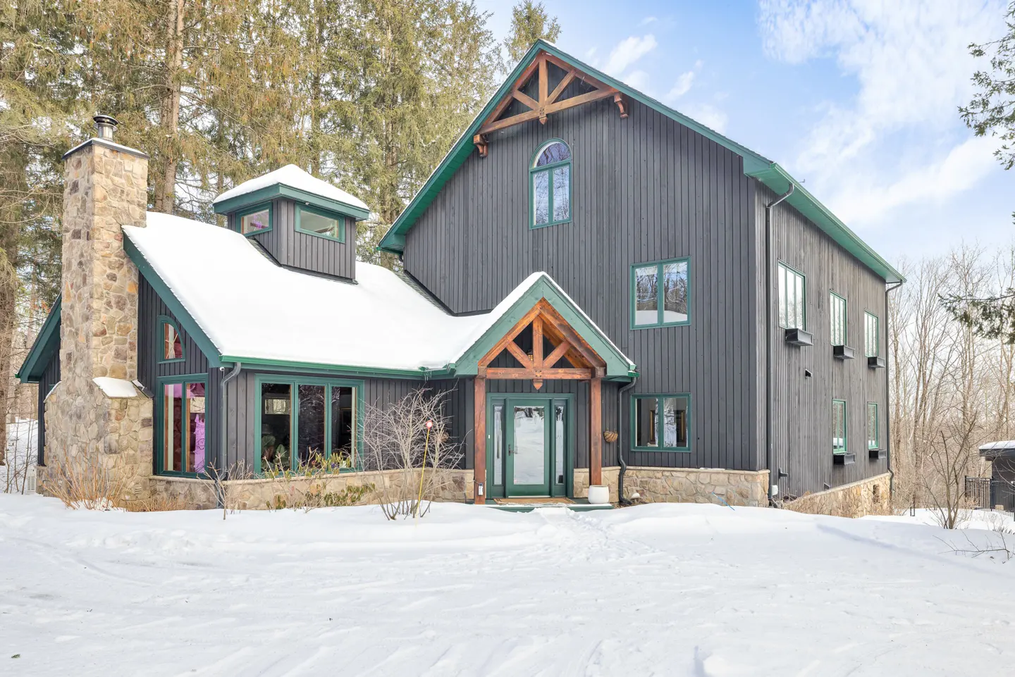 Exterior of a gray, two-story home with green trim and a stone chimney in a snowy landscape.