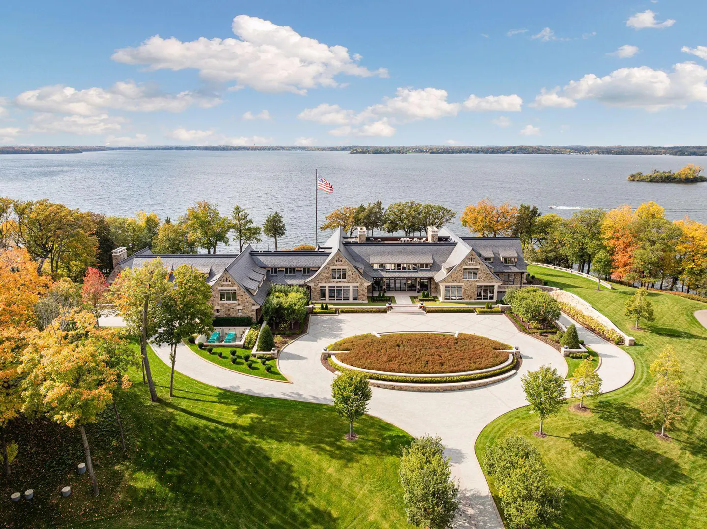 Aerial view of a large stone house with a circular driveway, green lawn, and lake in the background.