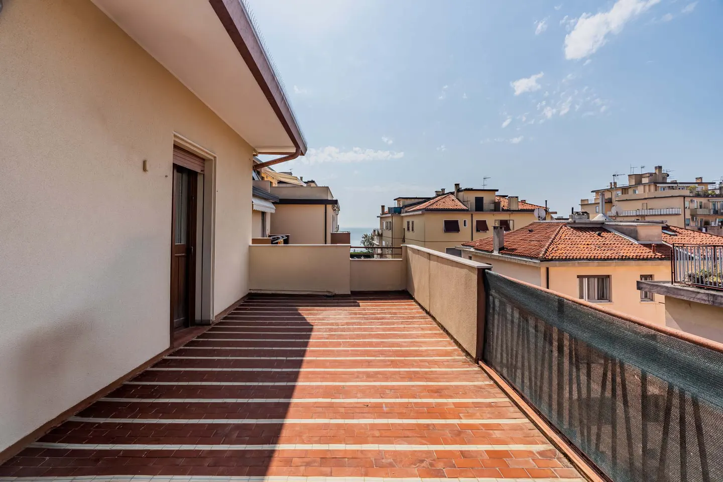 Wide balcony with red brick flooring and metal railing. Cityscape and blue sky visible in the background.