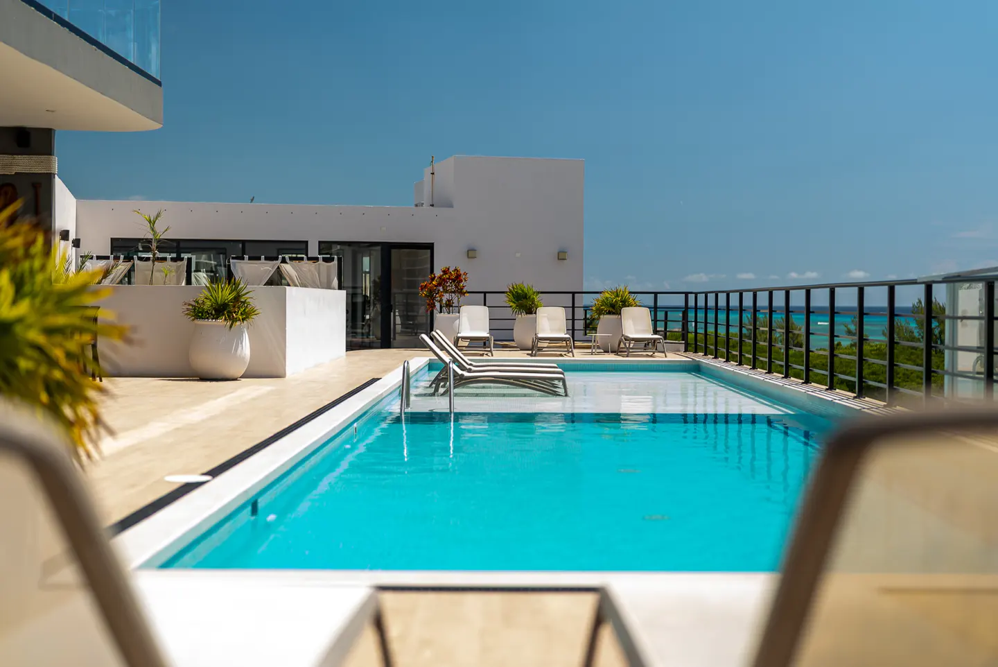 Rooftop pool with turquoise water, lounge chairs, and ocean view. White building and black railing. Sunny day.