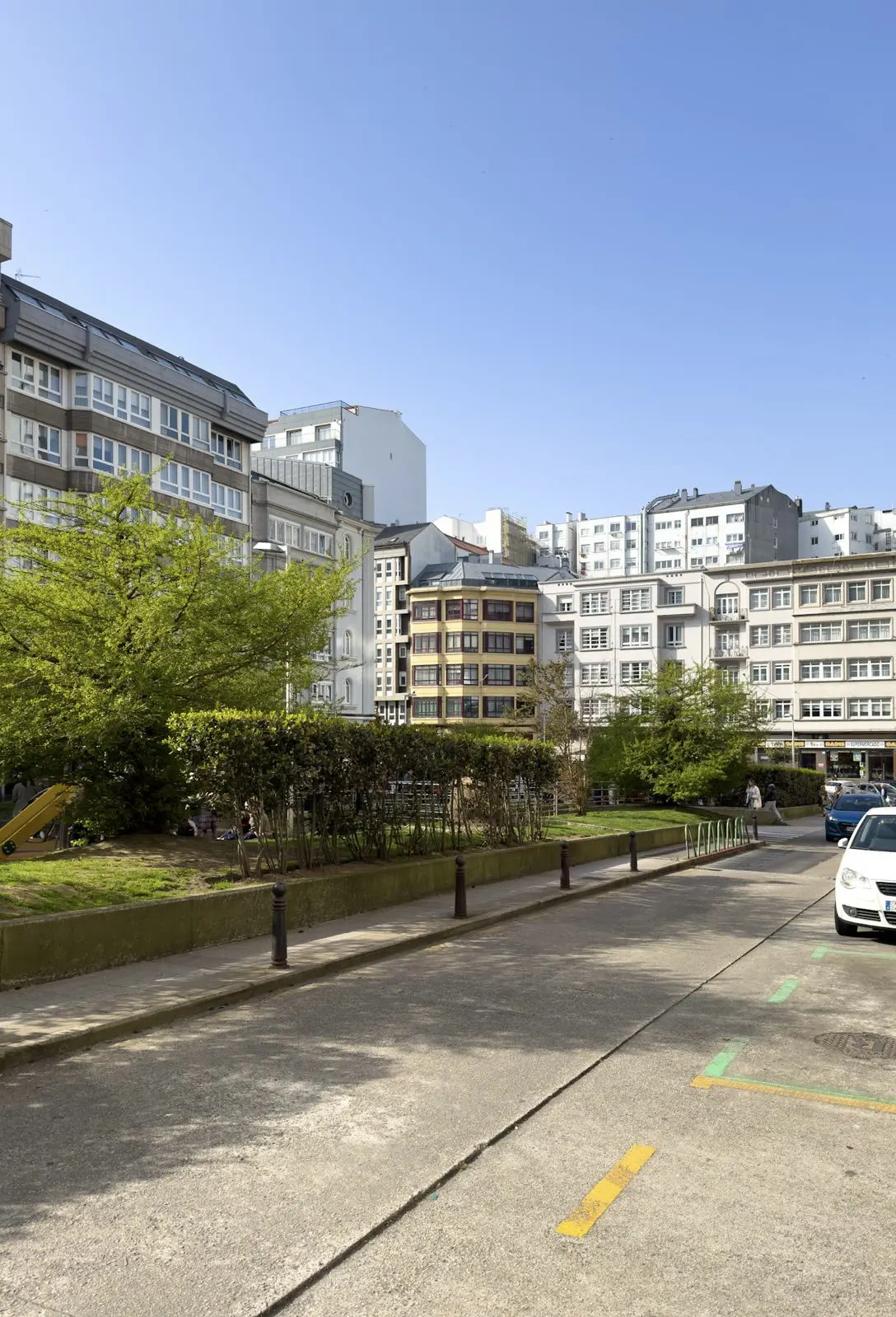 Cityscape view with apartment buildings, green trees, and a concrete parking lot in the foreground under a clear blue sky.