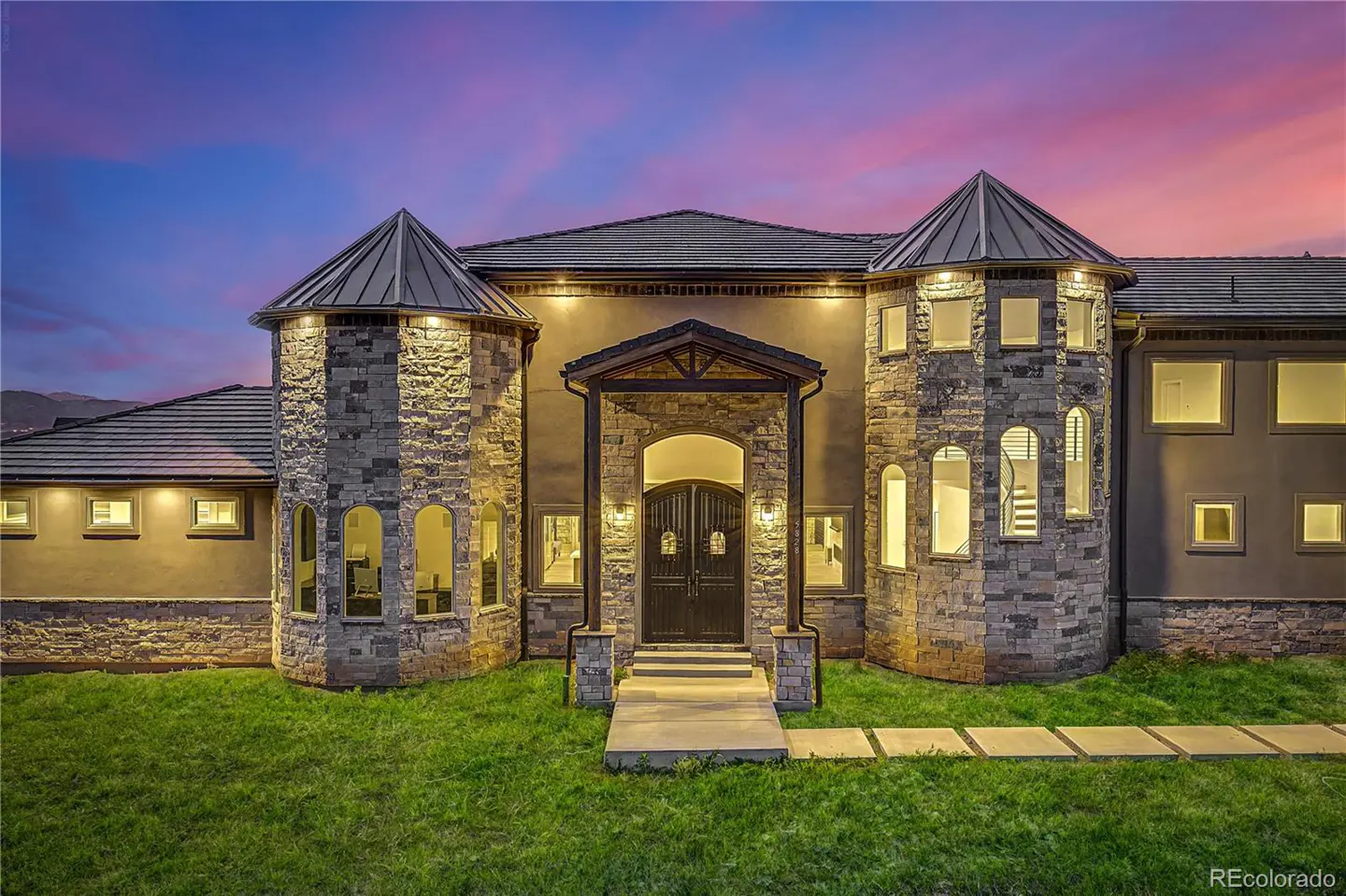 Luxury stone and stucco home with turrets and a covered entryway at dusk under a vibrant purple and orange sunset.