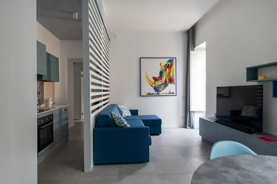A modern apartment interior with a blue sofa, TV, and colorful rhino art. A white slat wall separates the kitchen.