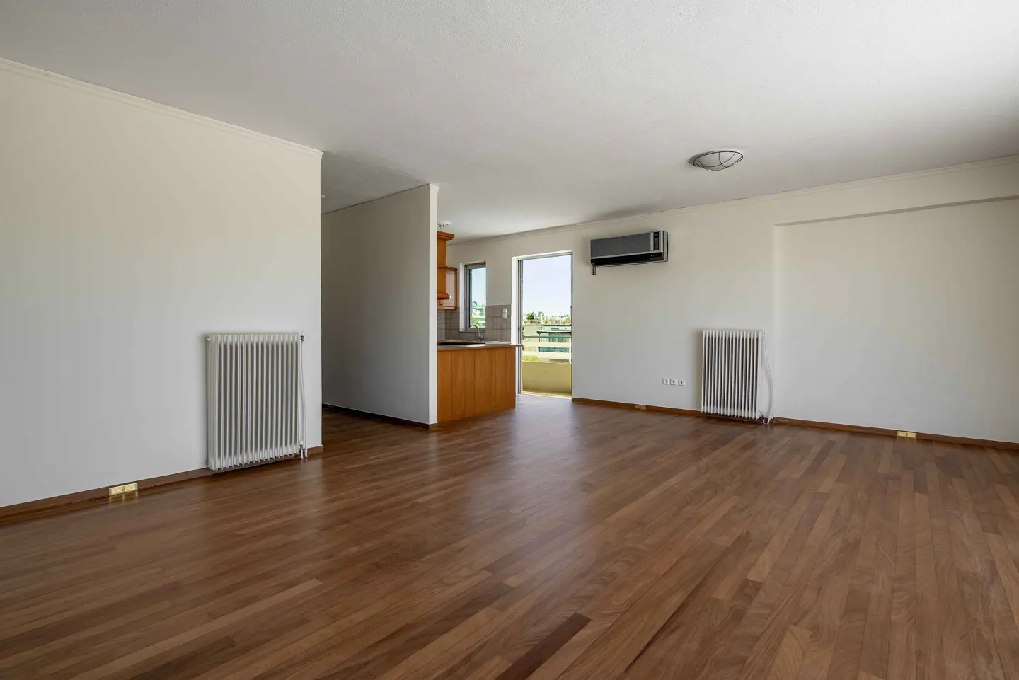Bright, empty apartment with hardwood floors, white walls, and a glimpse of the kitchen and balcony. Radiators and an AC unit are visible.