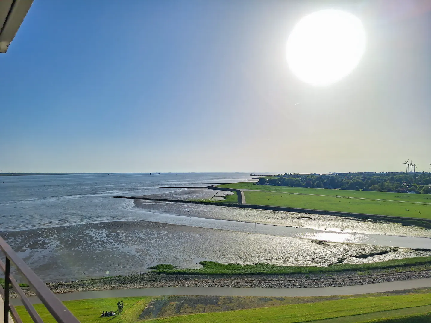 View from a balcony overlooking a sunny coastal landscape with mudflats, green fields, and wind turbines in the distance.