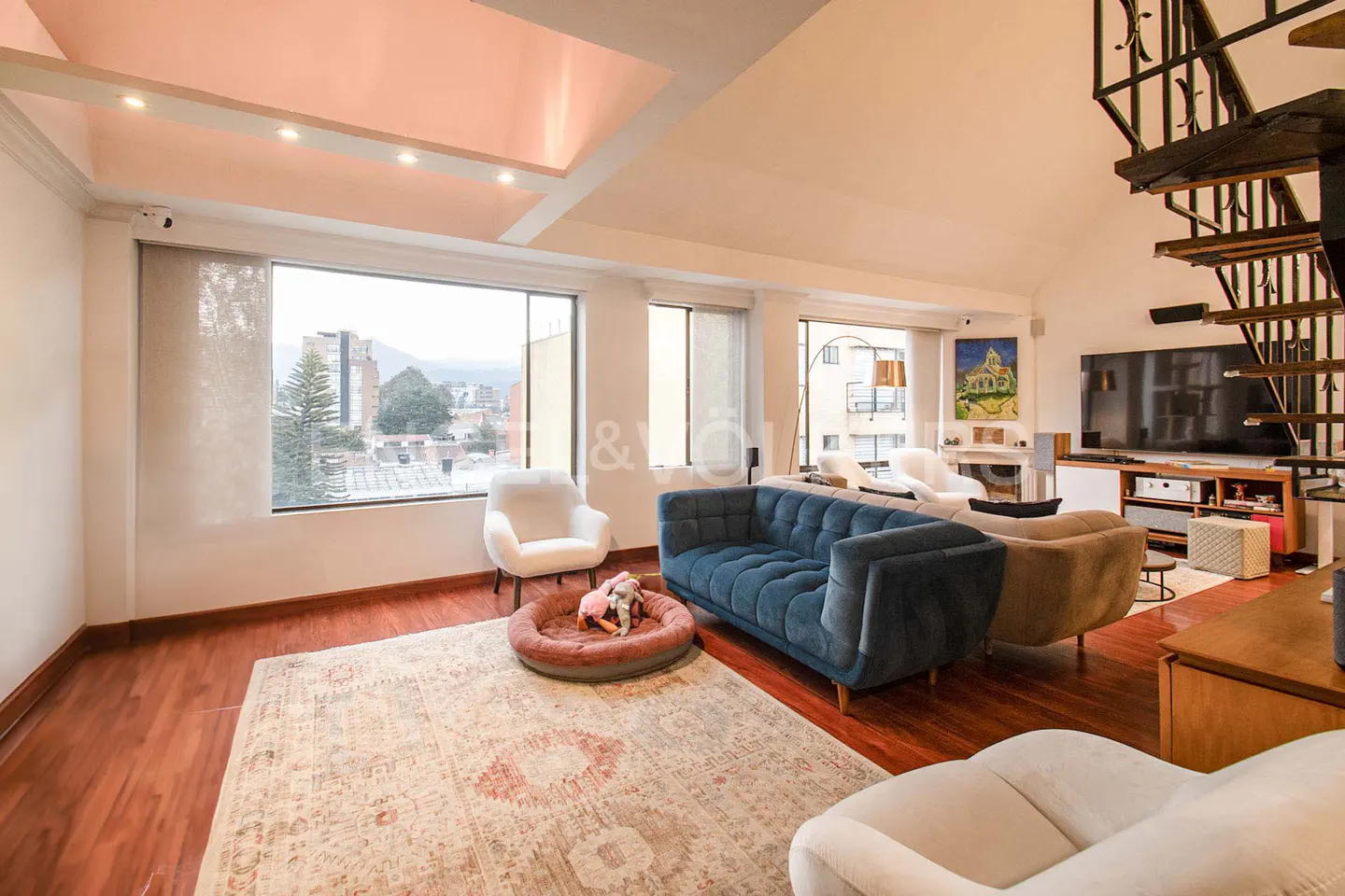Bright living room with a blue sofa, white chairs, and a patterned rug on a wood floor. A spiral staircase is visible.