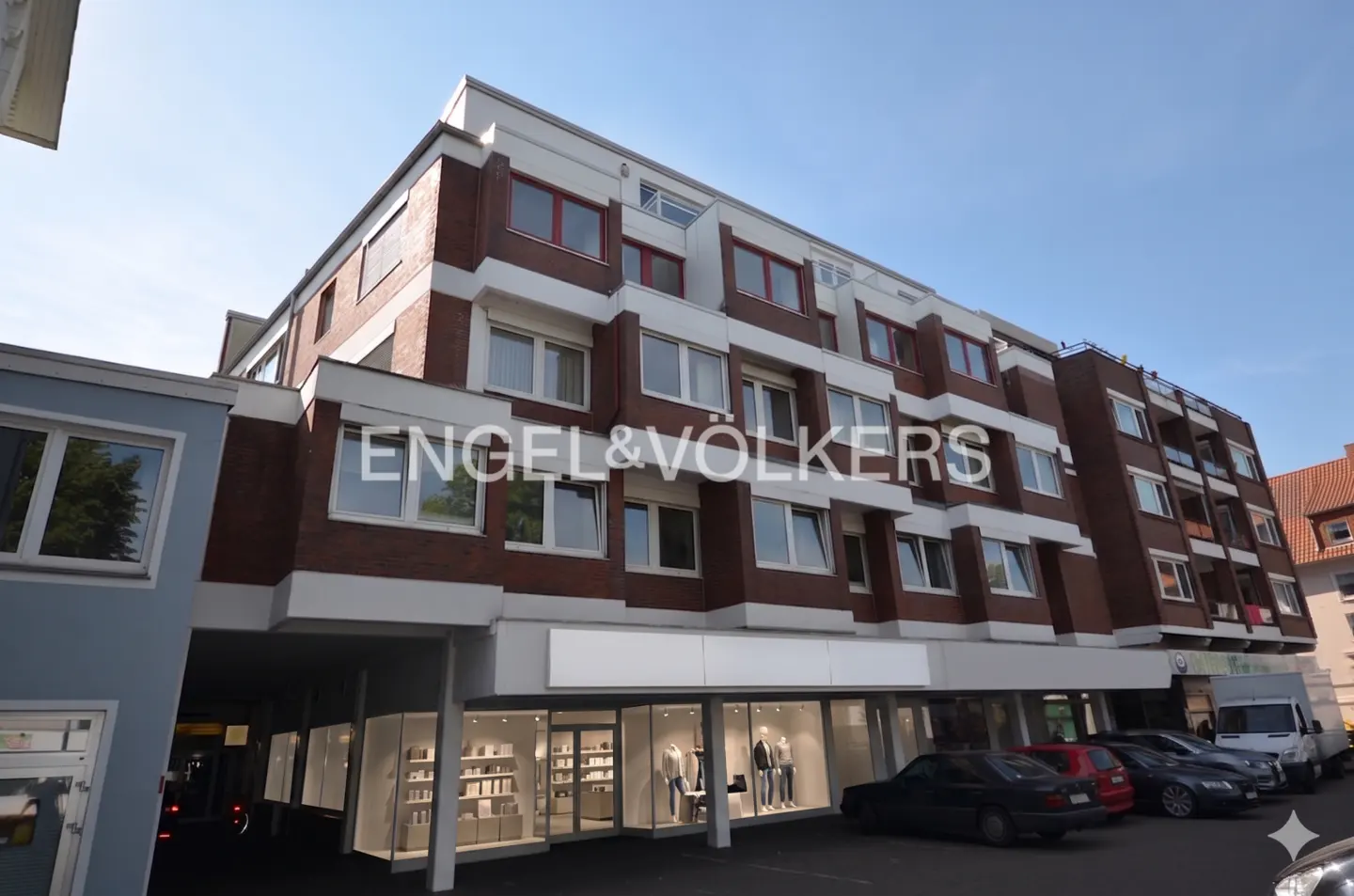 Exterior view of a multi-story brick building with white trim and large windows, cars parked in front. Engel & Völkers logo visible.