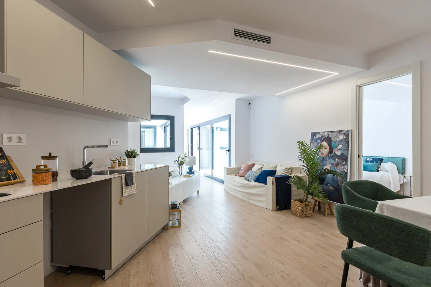 Open-concept living space with a kitchen island, white sofa with colorful pillows, and a large painting. Light wood floors and modern lighting.