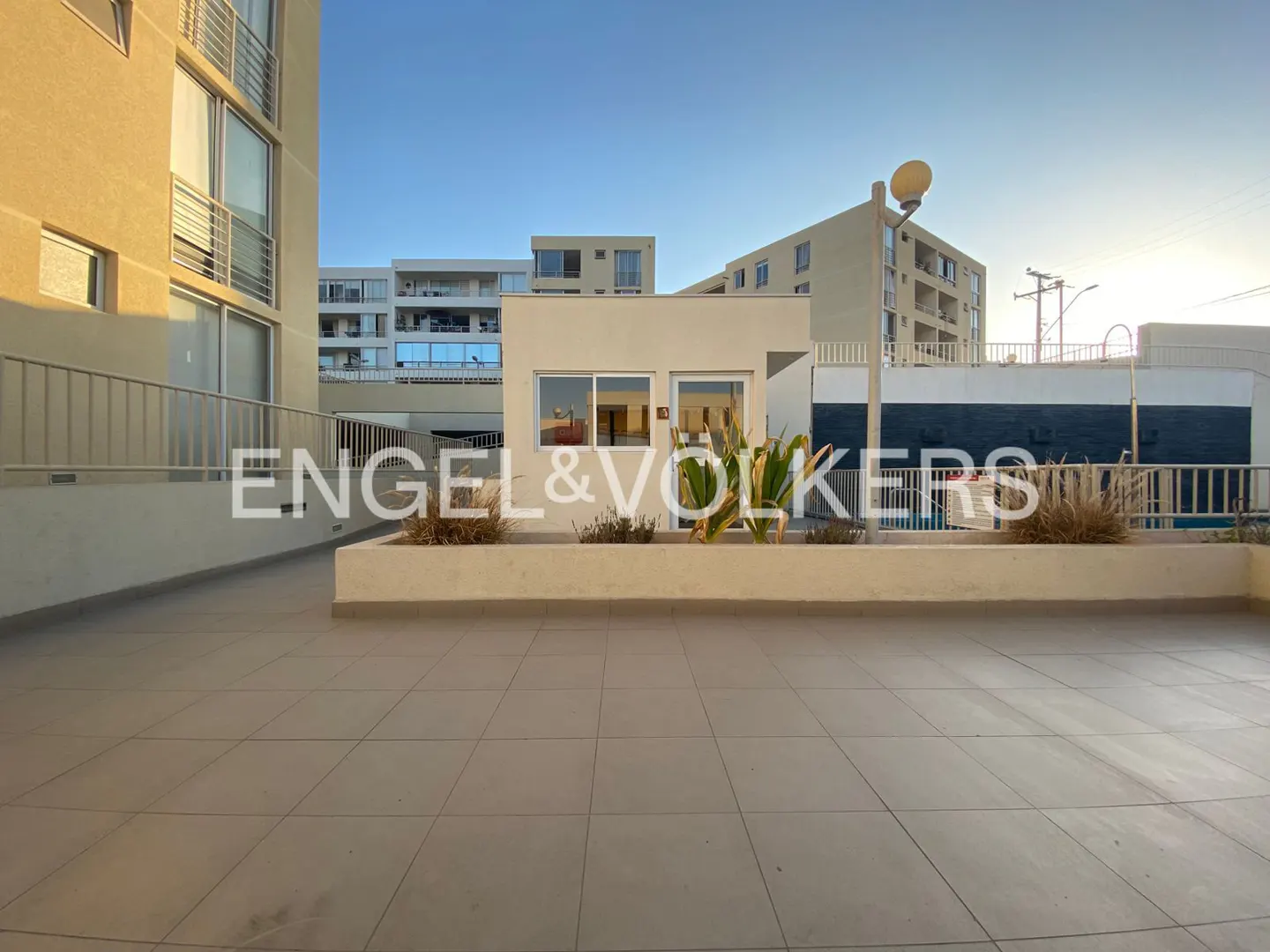 Exterior view of a tiled patio with a low wall and plants, buildings in the background, under a blue sky.