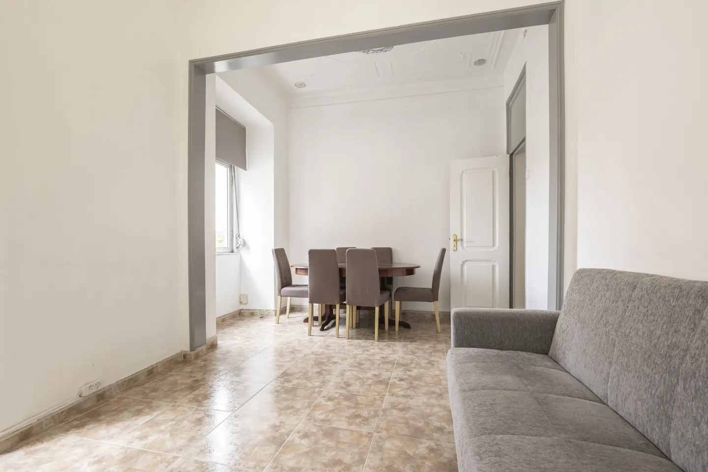 A bright room with a gray sofa, a dining table with six chairs, and a window with a gray blind.