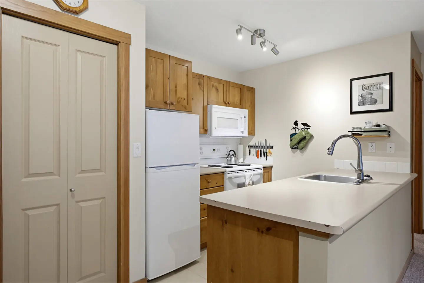 A bright kitchen with light wood cabinets, white appliances, and a breakfast bar with a sink. A coffee print hangs on the wall.