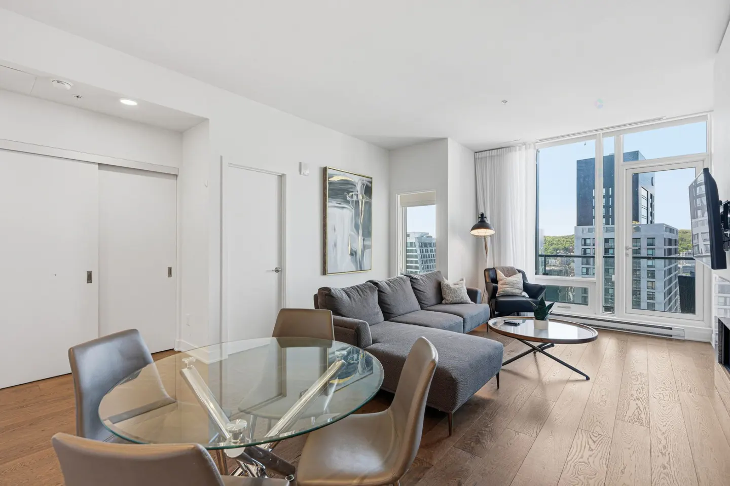 Bright living space with a gray sectional sofa, glass table, and city views from large windows. Hardwood floors and white walls.
