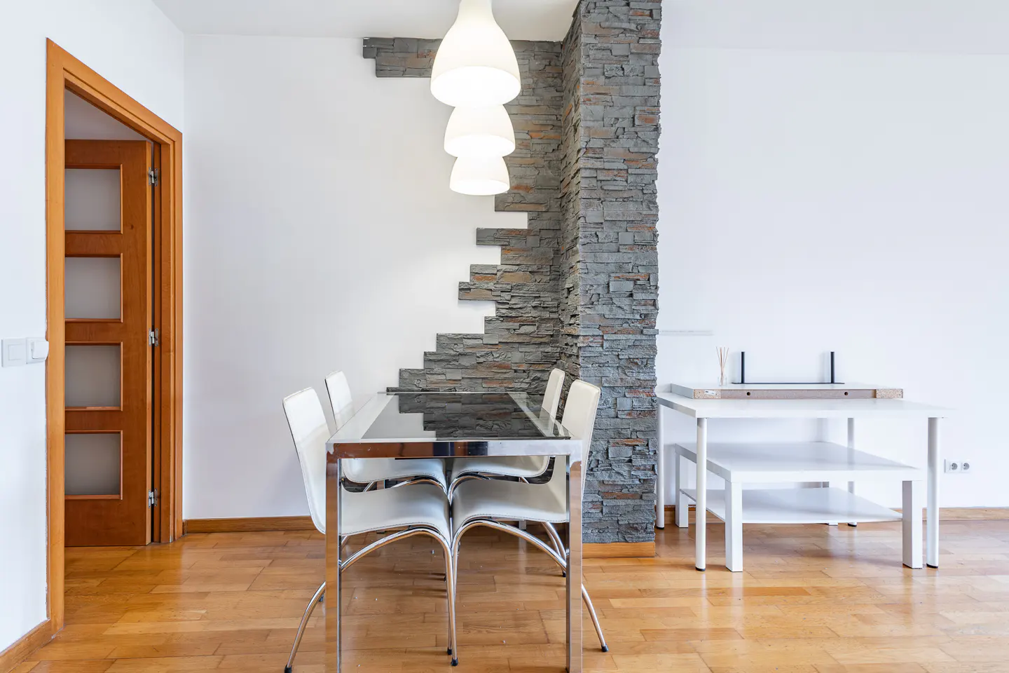 Modern dining area with a glass table, white chairs, and a decorative grey stone accent wall with three pendant lights.