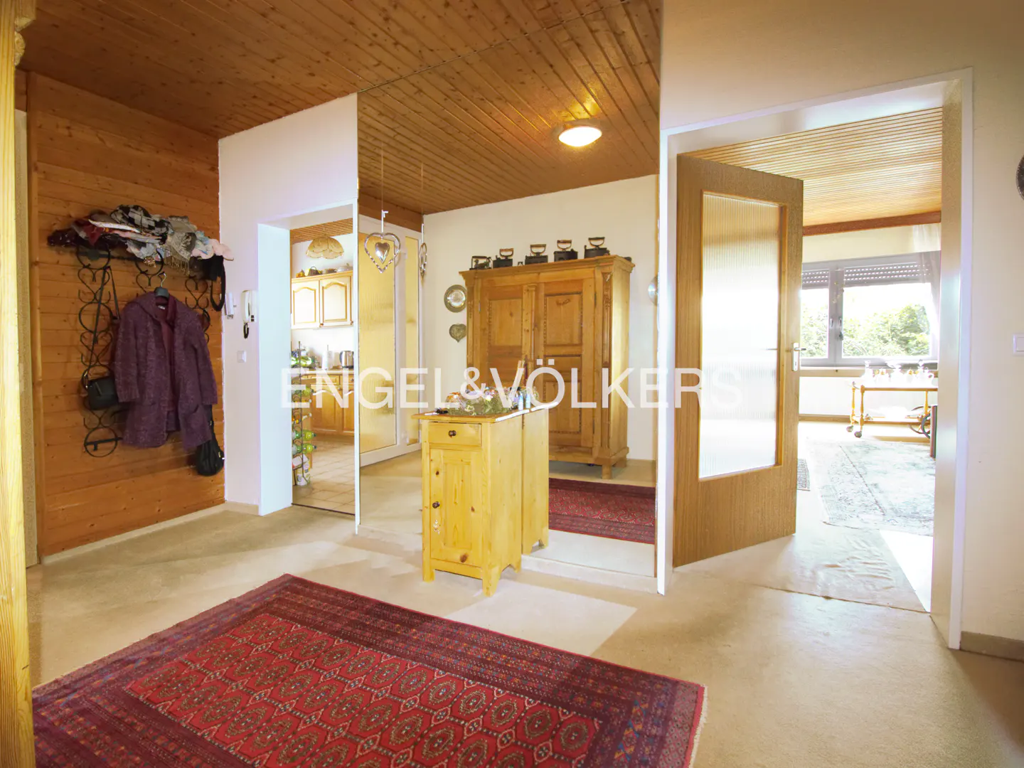 Entryway with a red patterned rug, wood-paneled ceiling, a coat rack with a purple jacket, and a wooden cabinet.