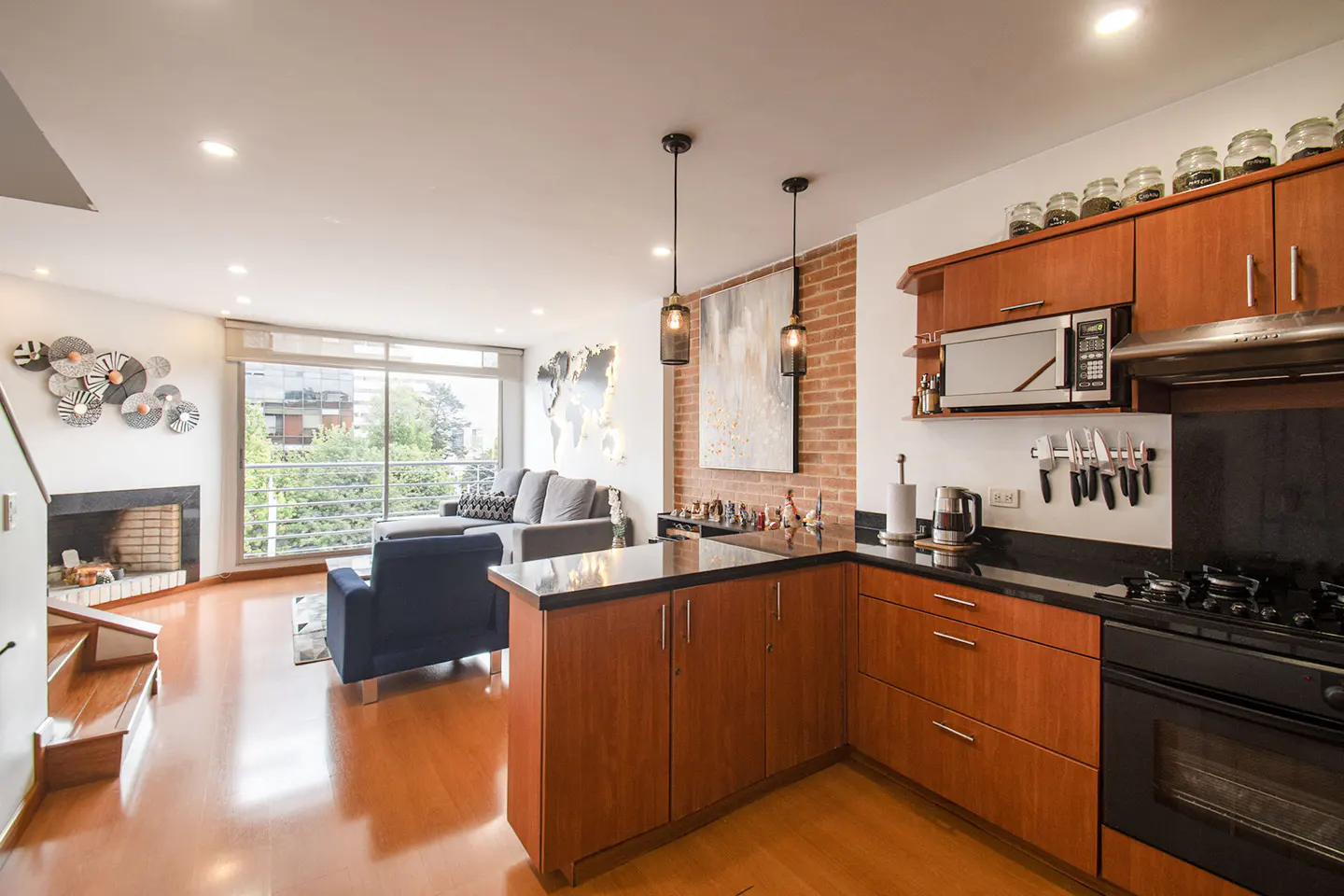 Open-concept living space with wood floors, featuring a kitchen with cherry cabinets and a living room with a fireplace and large window.