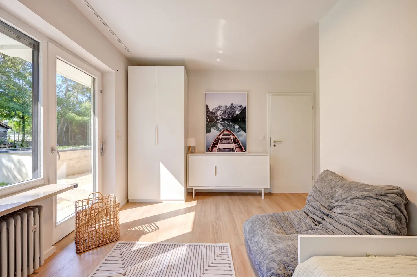 Bright room with wood floors, white walls, and natural light. Features a wardrobe, dresser with a boat picture, and a gray beanbag chair.
