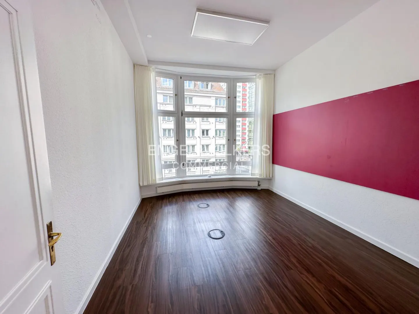 Empty room with dark wood floors, white walls with a horizontal red stripe, and a large window with white curtains.