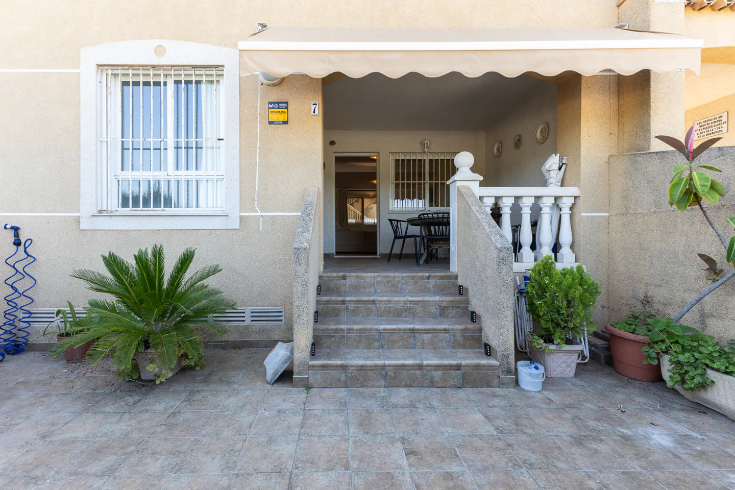 Exterior view of a beige house with a tiled patio, stone steps leading to a covered porch, and potted green plants.
