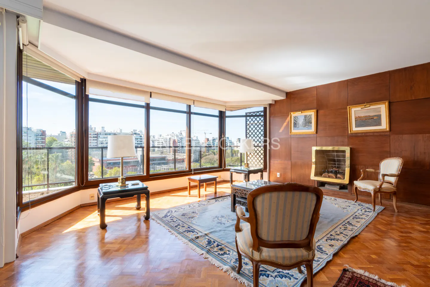 A bright living room with wood paneling, a fireplace, and a large window overlooking a city. A blue rug covers the parquet floor.