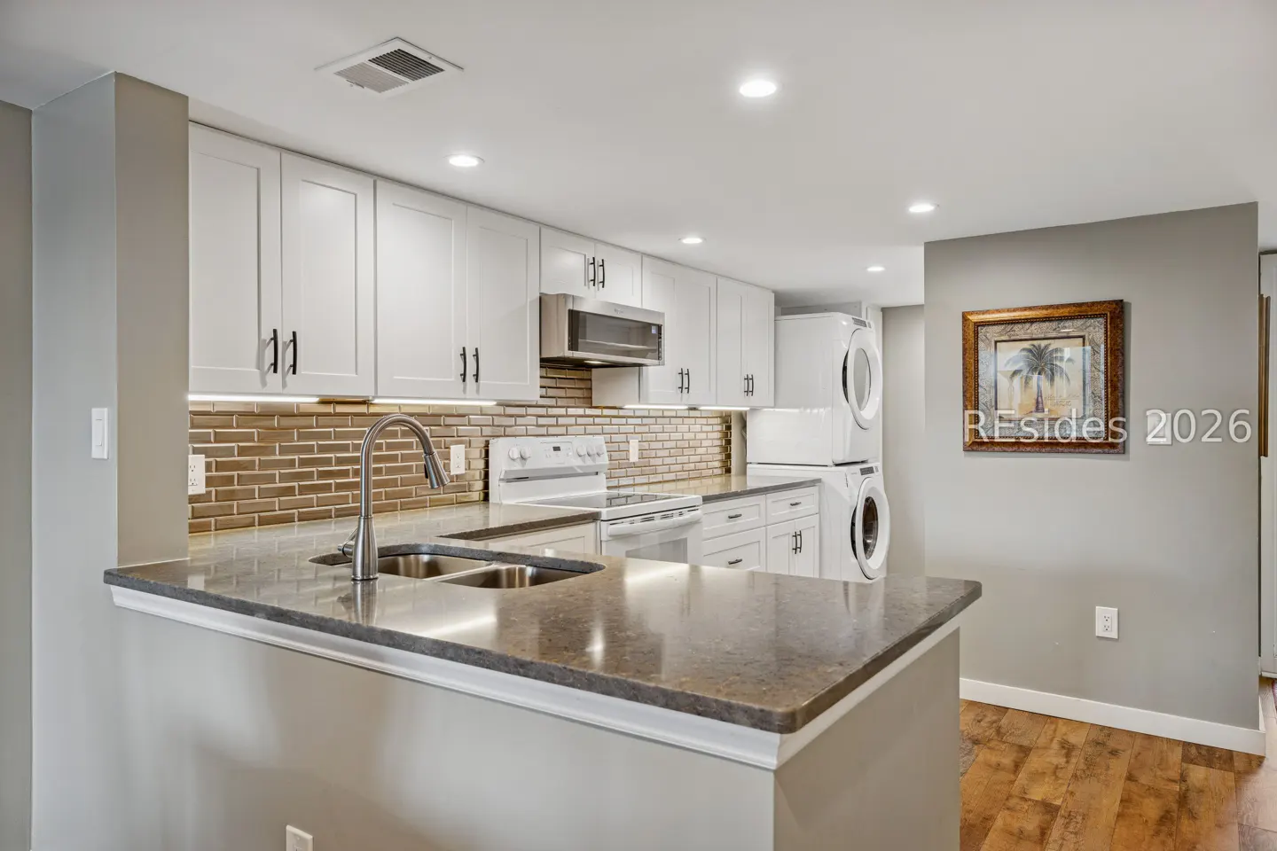 A kitchen with white cabinets, stainless steel appliances, and a brown tile backsplash. A stacked washer and dryer are visible in the background.